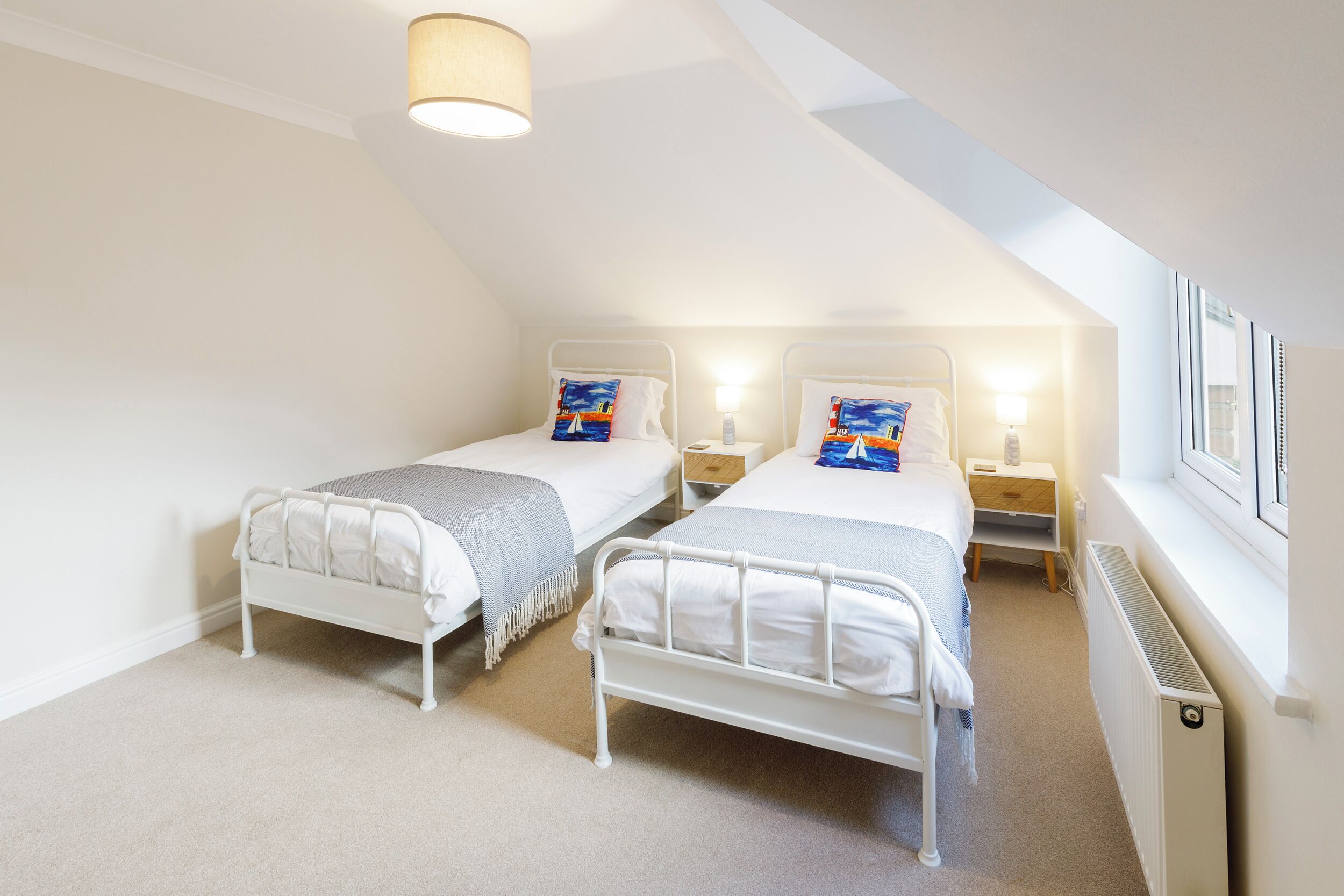 Un dormitorio con dos camas individuales, cada una con una mesita de noche y una lámpara. La habitación tiene un techo inclinado con un tragaluz y un radiador en la pared.