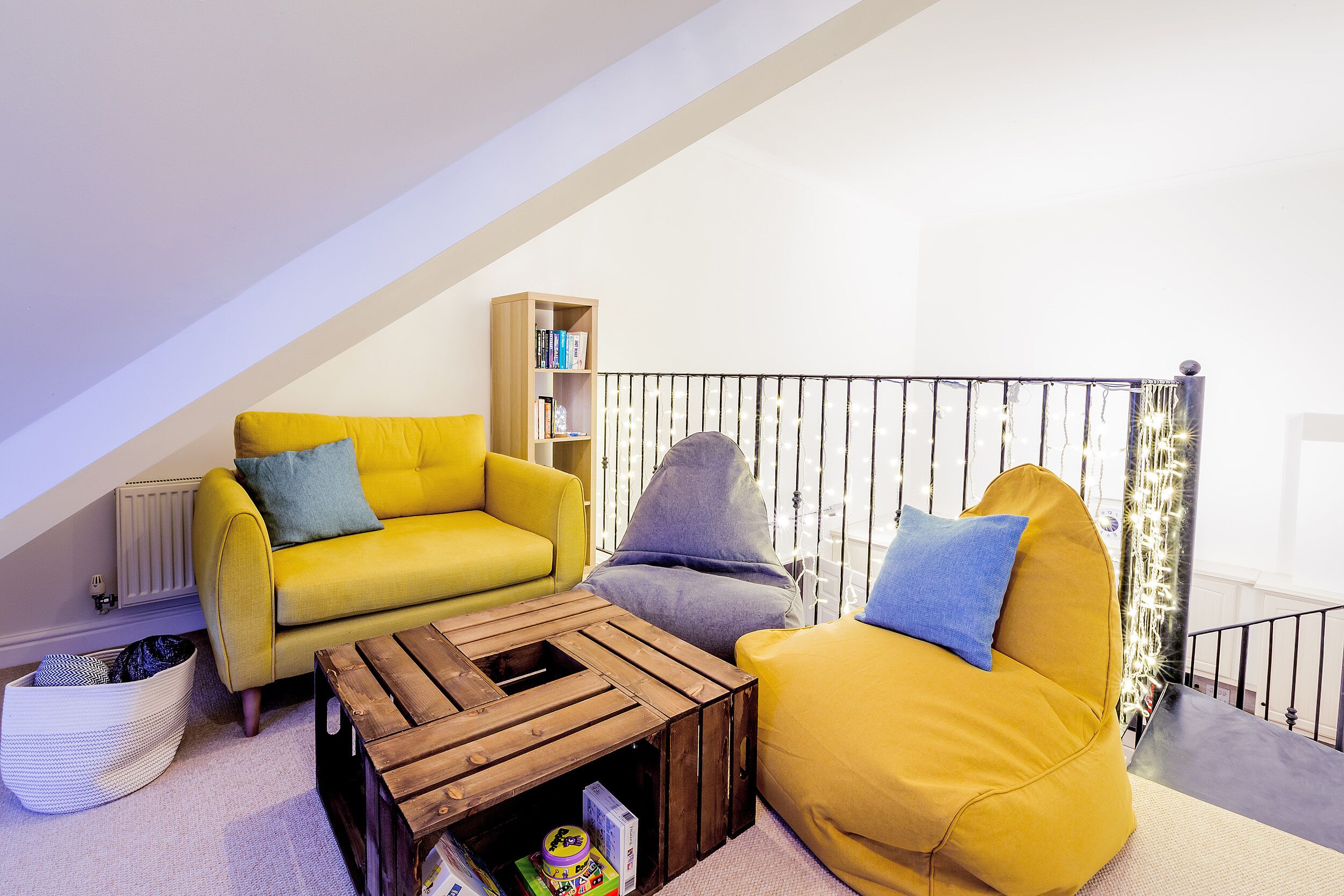 Un acogedor salón estilo loft con un sofá amarillo, un sillón tipo bean bag gris y una mesa de centro de madera. Una estantería está en la pared, y una barandilla negra separa la sala de estar del piso superior.