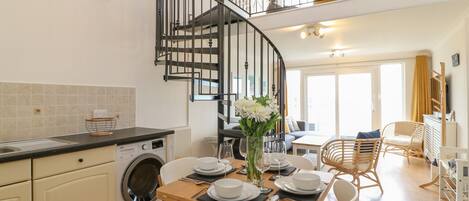 Una cocina moderna con lavadora integrada, un comedor con mesa para cuatro, y una escalera de caracol que sube a un nivel superior.