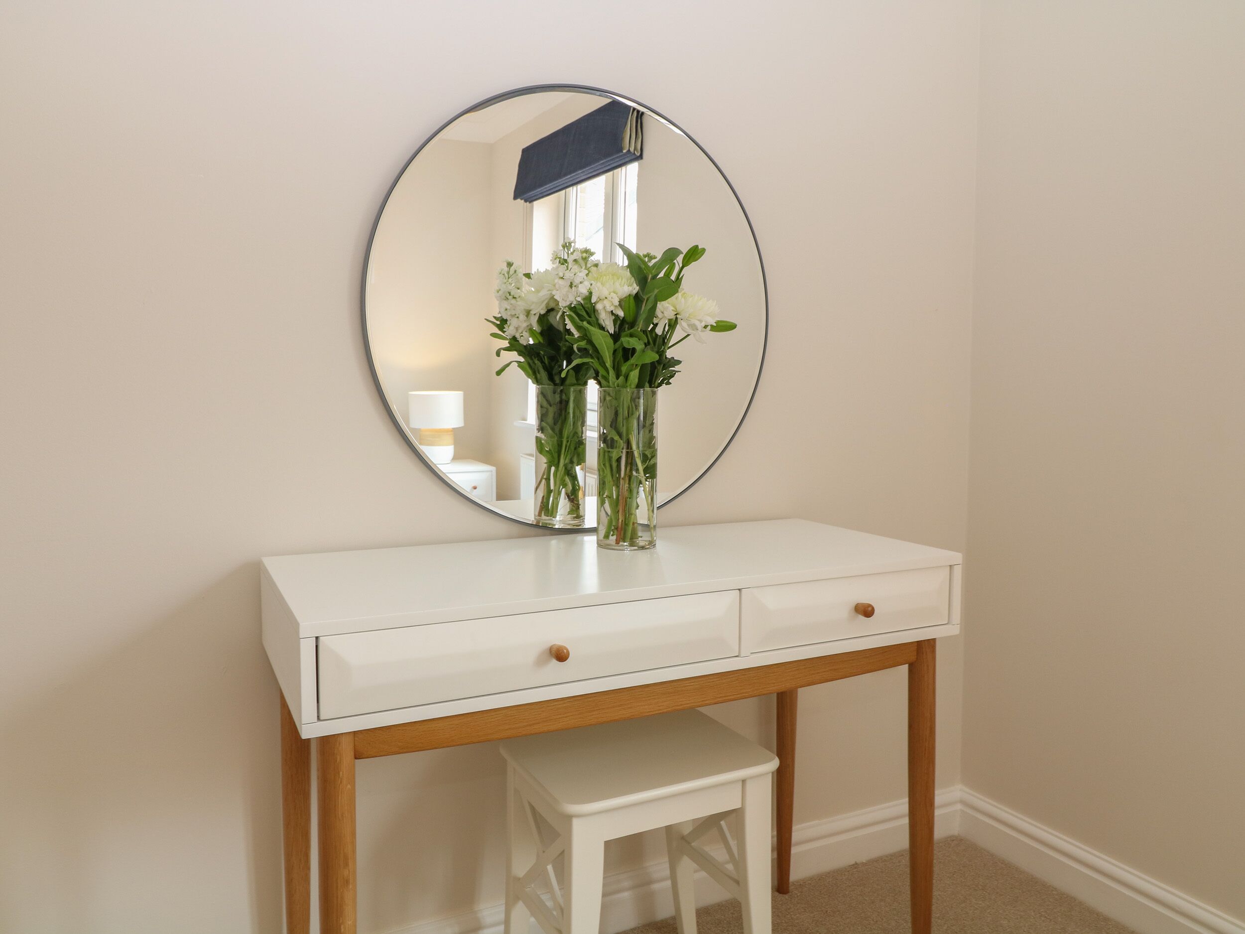 A white vanity with a round mirror, a small white stool, and a vase with flowers.