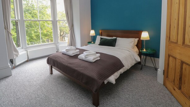 A neatly made bed with a brown bedspread and two pillows, a blue wall, and a wooden door. - Belvedere (Buxton)