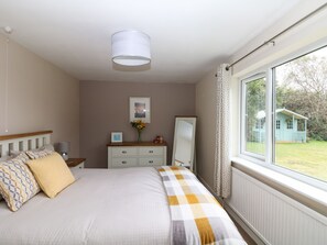 A bedroom with a bed, a bedside table, a mirror, a dresser, and a window with a view of a house. - Trevore Farmhouse (Truro)