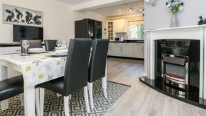 A modern kitchen with white cabinets, a black countertop, and a built-in microwave. There is a dining area with a white table and black chairs, and a fireplace with a black mantel. - Trevore Farmhouse (Truro)