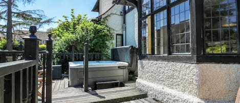 Una terraza de madera con un jacuzzi, un jardín cercado y un edificio con ventanas amplias.