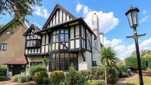 Una casa de estilo Tudor con un amplio ventanal saliente, un sendero de ladrillo y abundante vegetación circundante.