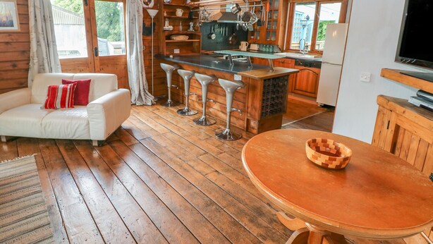 Ein gemütliches Blockhaushaus-Interieur mit Holzboden, einem weißen Sofa, einem runden Holztisch, einer Küchenzeile mit Bar und einem Fernseher.