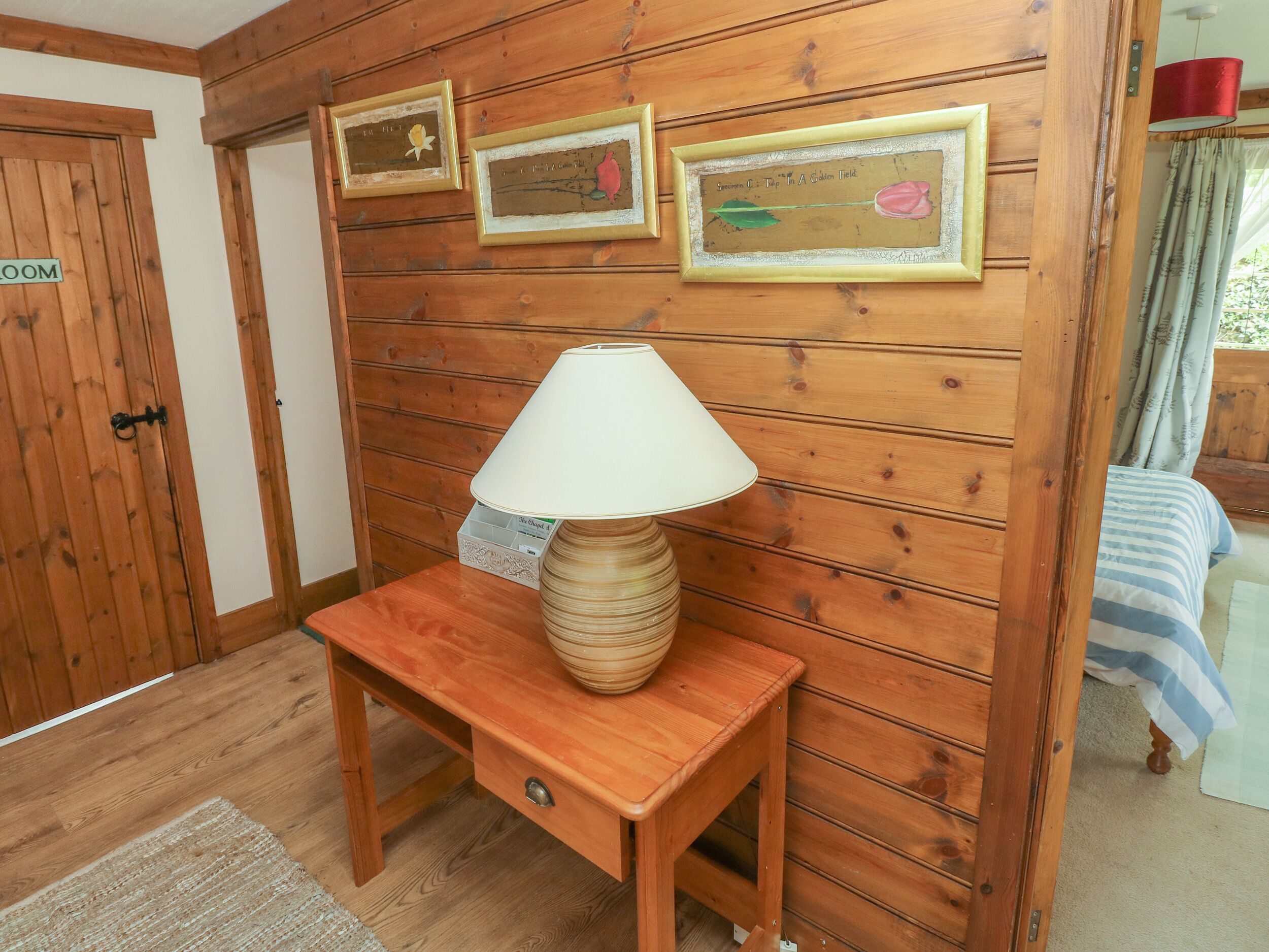 A wooden-paneled room with a wooden table, a lamp, and three framed artworks on the wall.
