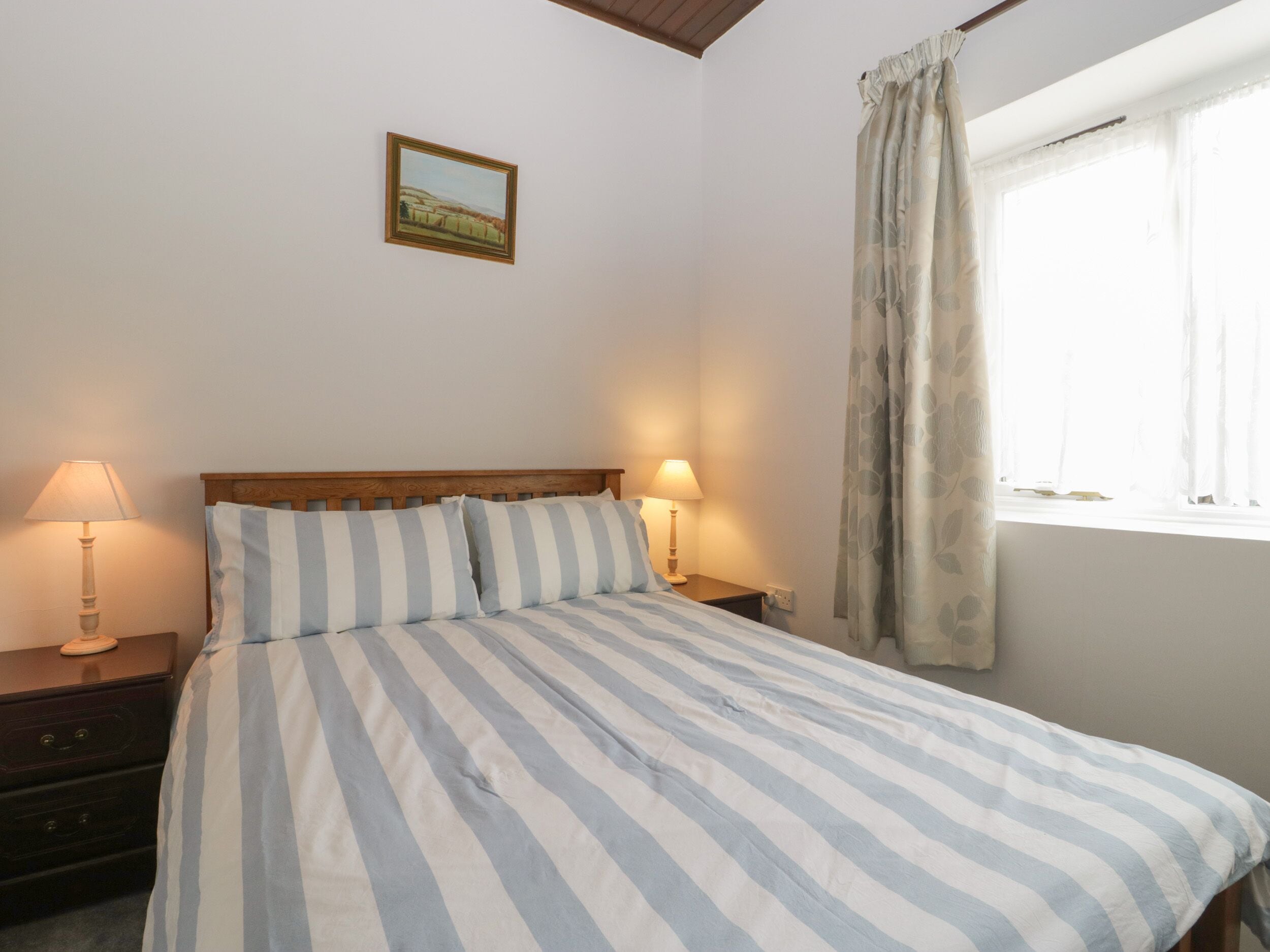 A bedroom with a bed, two bedside lamps, a nightstand, a window with curtains, and a framed picture on the wall.