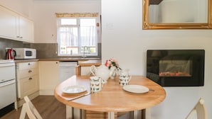A kitchen with wooden cabinets, a microwave, a sink, and a window with curtains. There is a dining table with chairs and a vase of flowers. - Stable Cottage (Taunton)