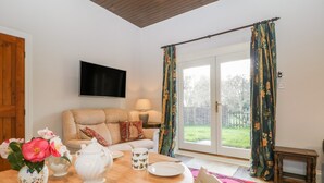 A cozy living room with a sofa, a TV mounted on the wall, and a dining area with a table set for two. - Stable Cottage (Taunton)