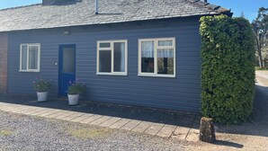 A small, single-story house with a blue exterior, a chimney, and two windows with white frames. - Stable Cottage (Taunton)