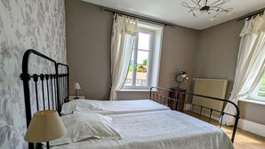 A bedroom with two beds, a bedside table, a lamp, a window with curtains, a radiator, and a decorative wall piece. - Charming 5-Bedroom House with Large Garden, Terrace, and Authentic Decor in Le Val-d'Ajol (Le Val-d'Ajol)
