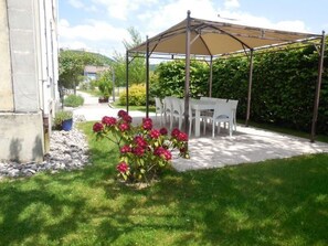 A covered outdoor seating area with a table and chairs, surrounded by a garden with red flowers and a hedge. - Charming 5-Bedroom House with Large Garden, Terrace, and Authentic Decor in Le Val-d'Ajol (Le Val-d'Ajol)