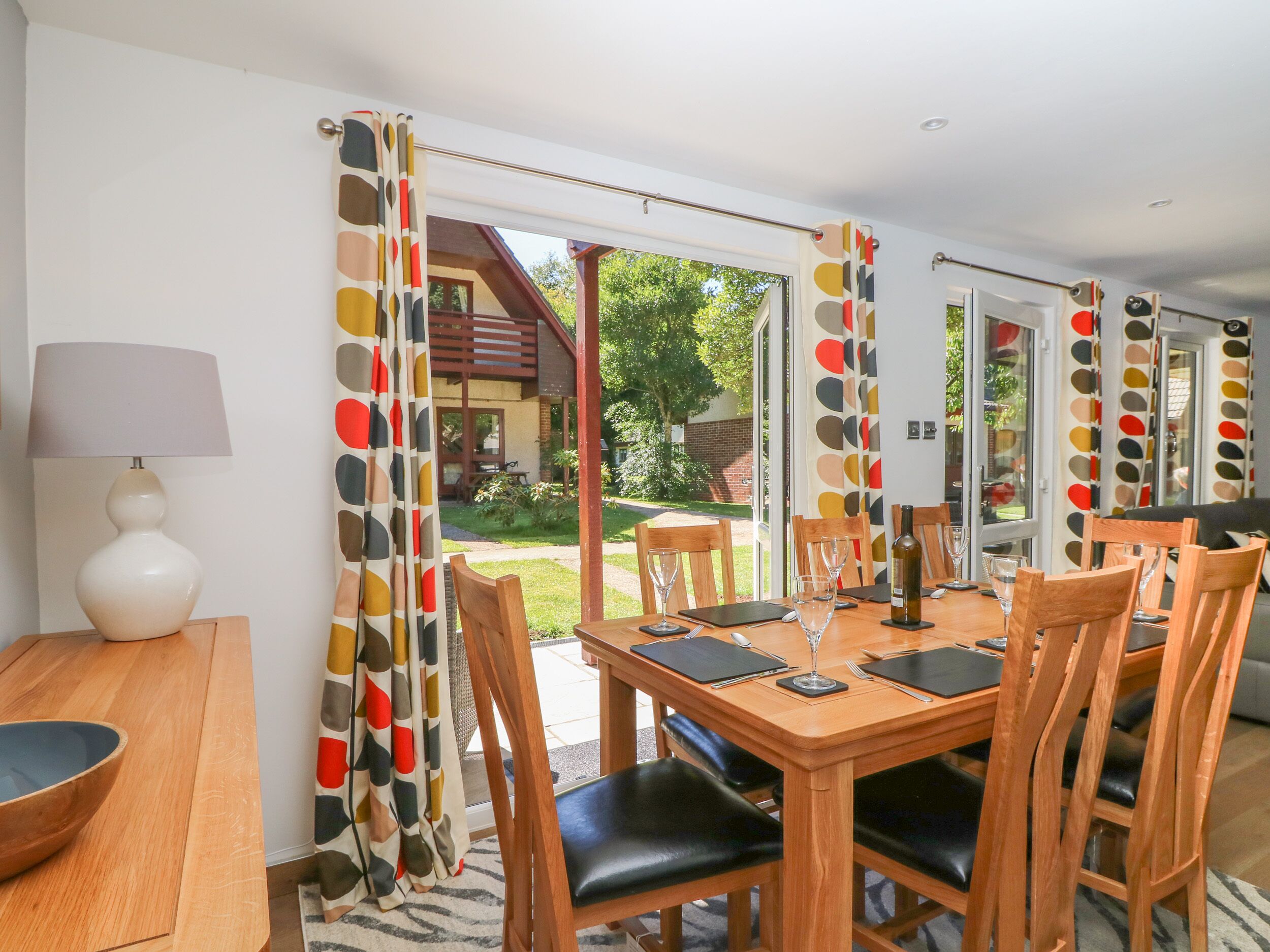 Ein Essbereich mit Holztisch und Stühlen, einer Lampe und Blick auf ein Gebäude und Garten durch große Fenster.