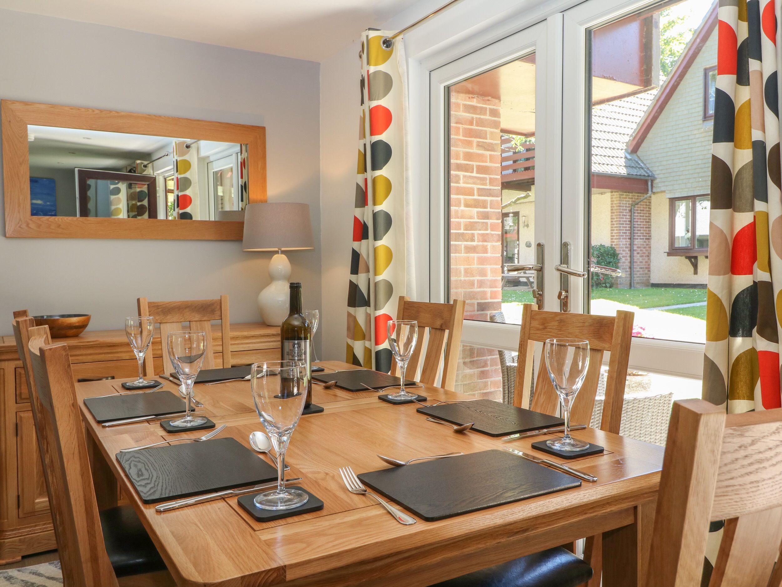 A dining area with a wooden table set for six, including wine glasses and cutlery. A bottle of wine is on the table, and there is a mirror on the wall.