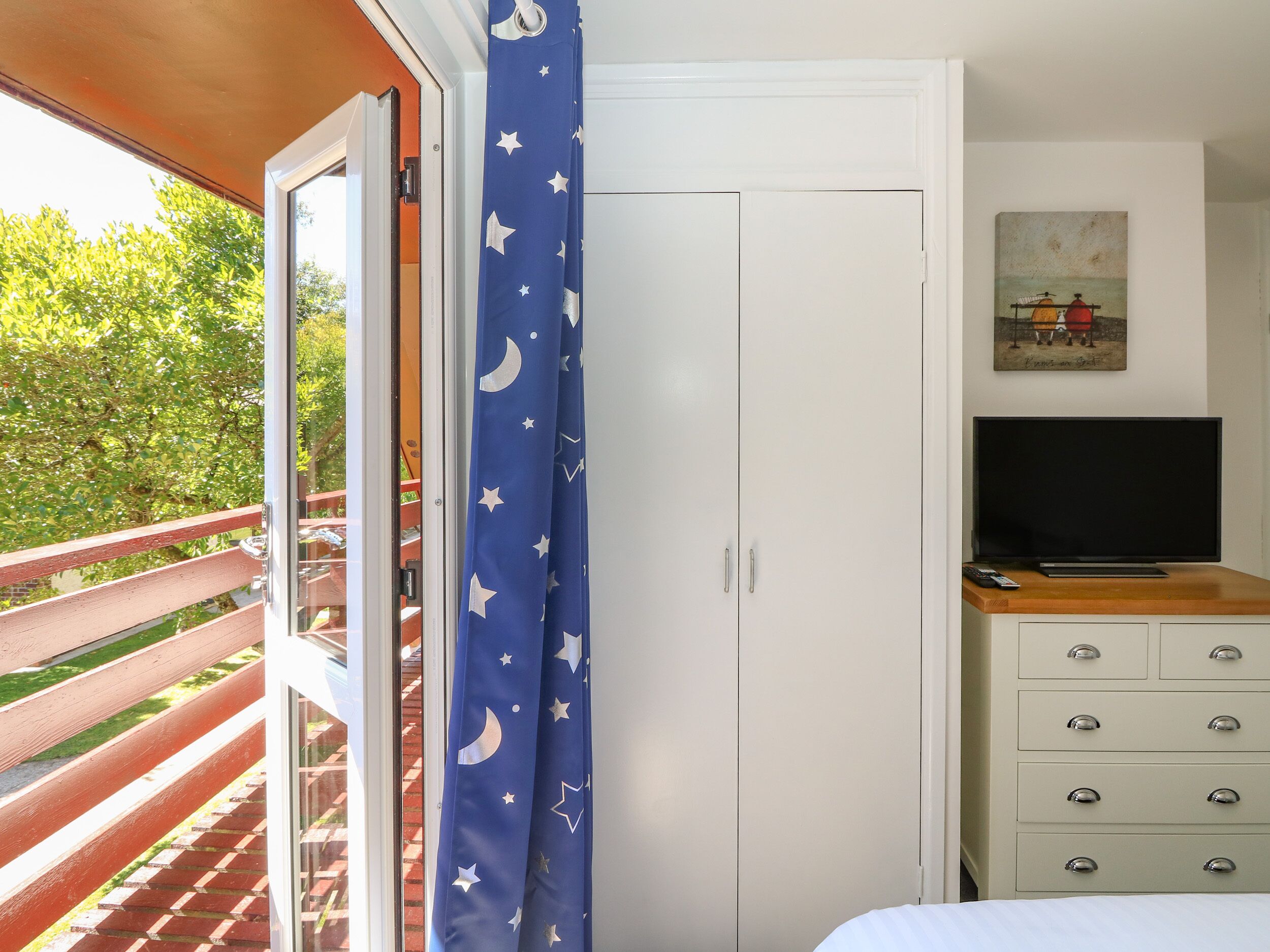 Un dormitorio con una puerta corrediza de vidrio que da a un balcón, un armario blanco, un televisor sobre un tocador de madera y un cuadro en la pared.