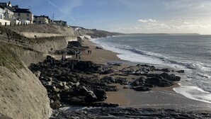 Un paysage côtier avec une plage rocheuse, une falaise sur laquelle se trouvent des bâtiments, et l’océan.