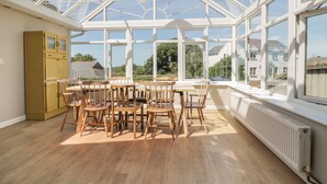A conservatory with a table and chairs, large windows, and a view of houses. - Auverne (Bude)