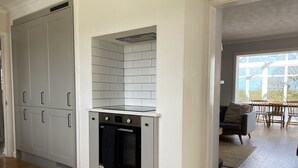 A compact kitchen with a built-in oven and a small stovetop, adjacent to a dining area with a view through large windows. - Auverne (Bude)