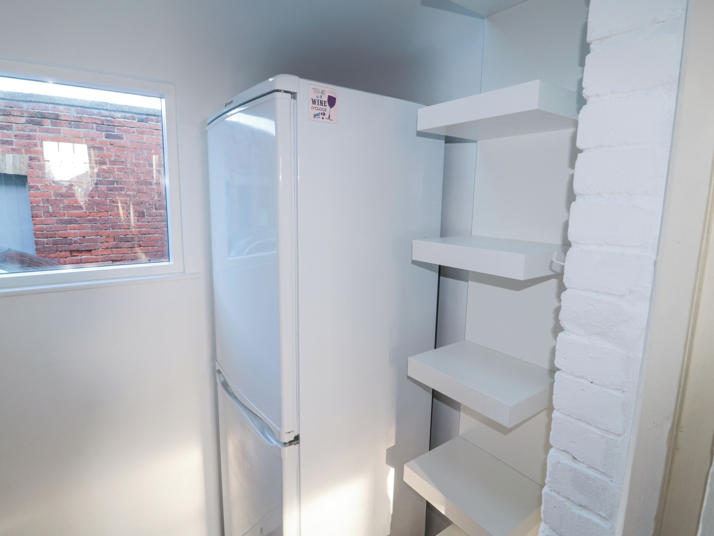 A compact kitchen with a refrigerator, a window with a view of a brick wall, and white shelves.