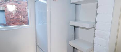 A compact kitchen with a refrigerator, a window with a view of a brick wall, and white shelves.