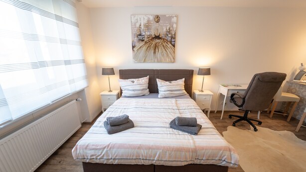A bedroom with a large bed, two bedside tables with lamps, a desk, a chair, and a large window with blinds. - Ferienwohnung Anke - Appartement 5 (Heinsberg)