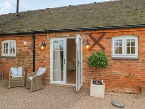 A brick building with a white door, two wicker chairs, a potted plant, and a gravel driveway. - March Hare (Ashbourne)