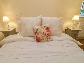 A neatly made bed with white bedding and decorative pillows, including a floral one. There are two bedside lamps on nightstands, and a window with a view of greenery outside.