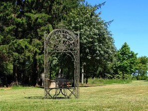 A gazebo with a metal frame and a bench inside, surrounded by a grassy area and trees.