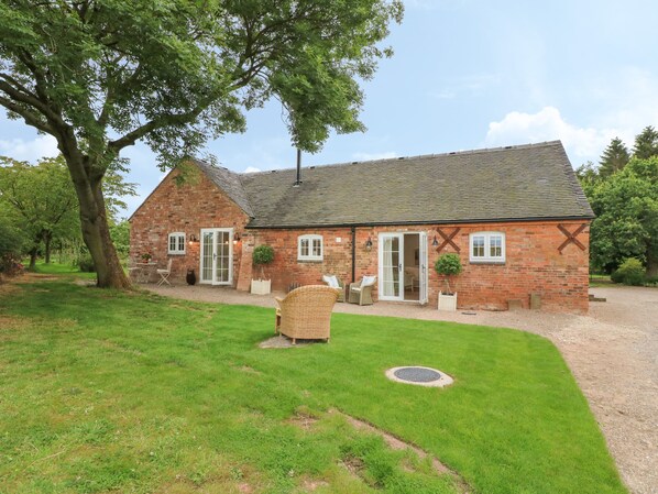 A brick house with a large tree in the front yard, a gravel driveway, and a small lawn area with outdoor furniture.