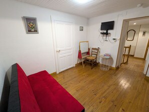 Una habitación con piso de madera, un sofá rojo, una silla de madera, una mesita con libros, una puerta blanca, una pared blanca con un cuadro enmarcado y un televisor montado en la pared.