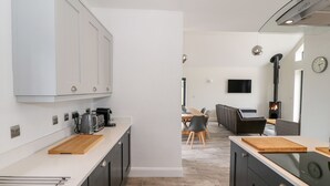Et moderne køkken med mørke skabe, en hvid bordplade og et indbygget komfur. Der er et spiseområde med bord og stole, samt en stue med sofa og fjernsyn.