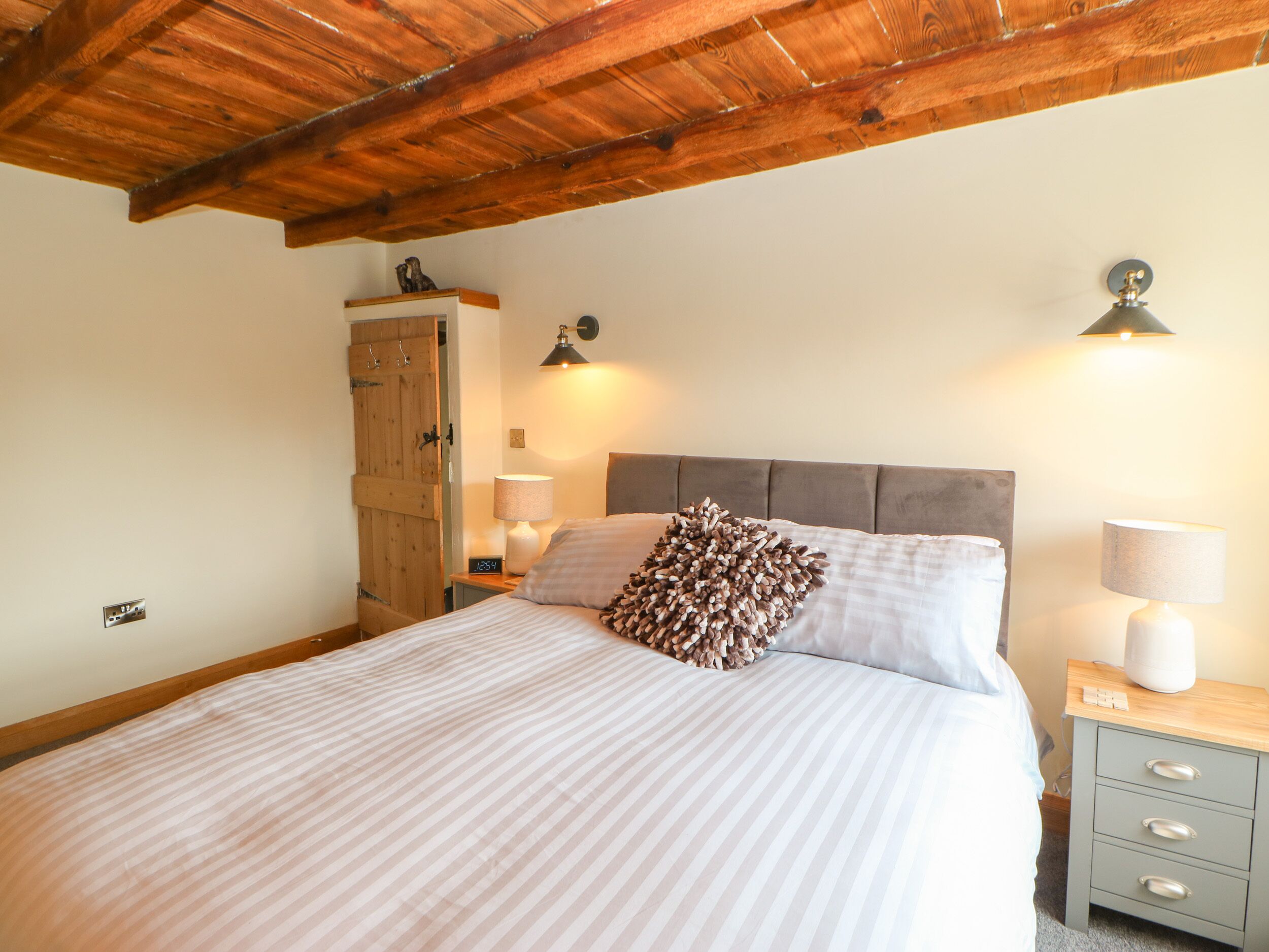 A bedroom with a wooden ceiling, a bed with striped bedding, two bedside lamps, a nightstand, and a wooden wardrobe.