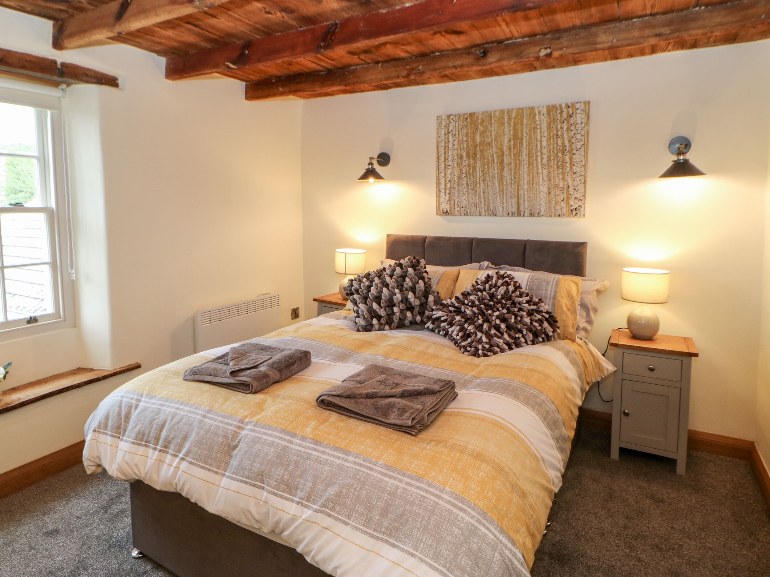 Quarto com uma cama, duas mesas de cabeceira, um radiador, uma janela e um vigamento de madeira no teto.