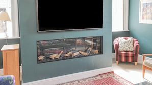 A living room with a flat-screen TV mounted on the wall, a fireplace with logs, a striped armchair, and a wooden side table. - Jubilee House (Alnwick)