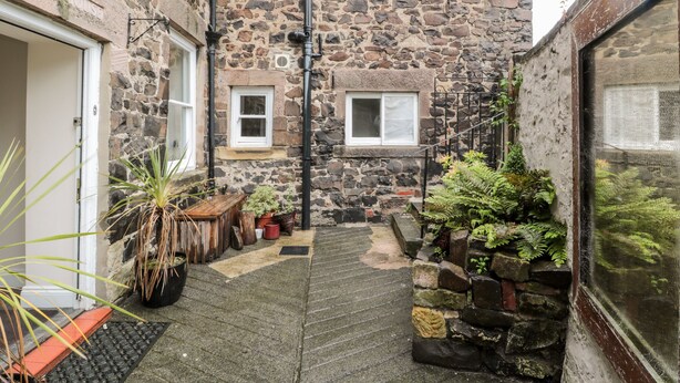 A stone building with a narrow courtyard, potted plants, and a wooden bench. - Jubilee House (Alnwick)