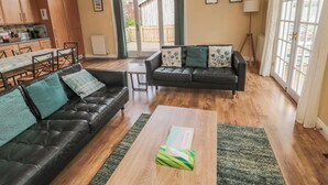 A spacious living area with a black leather sofa, a wooden coffee table, and a dining table with chairs. There is a kitchen in the background, a sliding glass door leading to an outdoor area, and a large window providing natural light. - The Shieling (Morpeth)