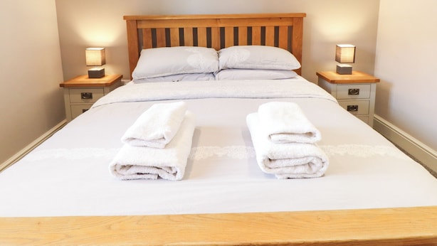 Una cama bien hecha con dos toallas blancas dobladas al pie, un cabecero de madera y dos lámparas de noche sobre mesitas a juego.