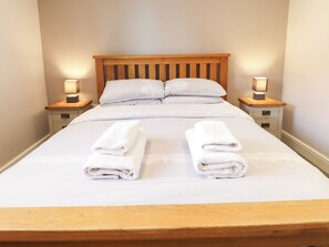 Una cama bien hecha con dos toallas blancas dobladas al pie, un cabecero de madera y dos lámparas de noche sobre mesitas a juego.
