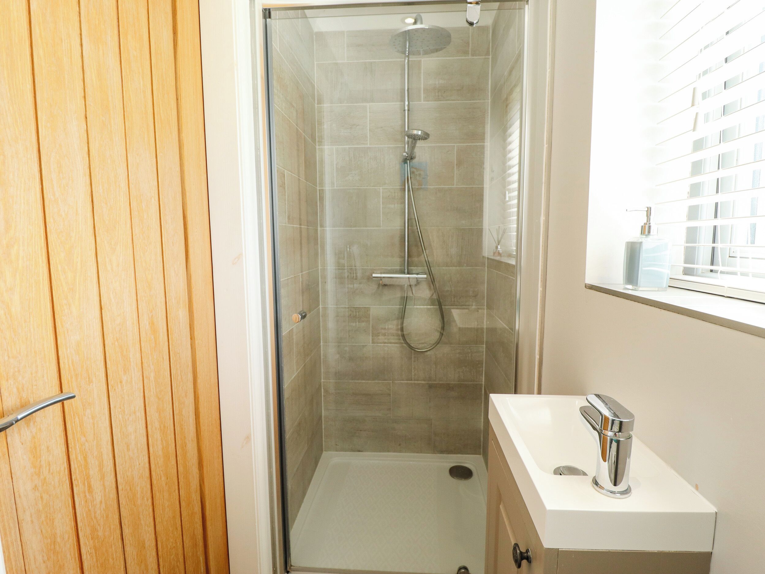 A bathroom with a glass-enclosed shower, a white sink with a chrome faucet, and a window with blinds.