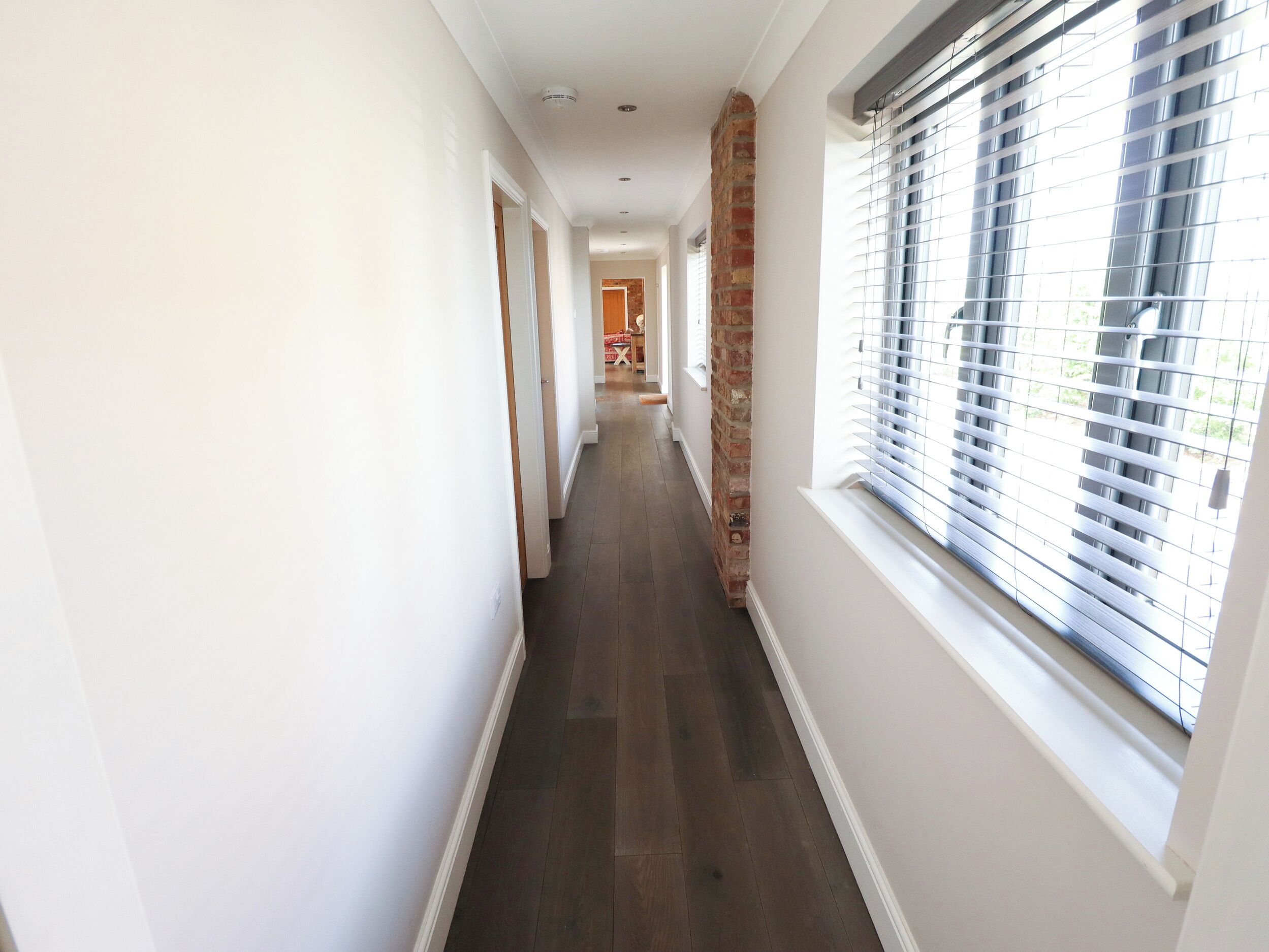 A long, narrow hallway with a wooden floor, white walls, and a brick column.