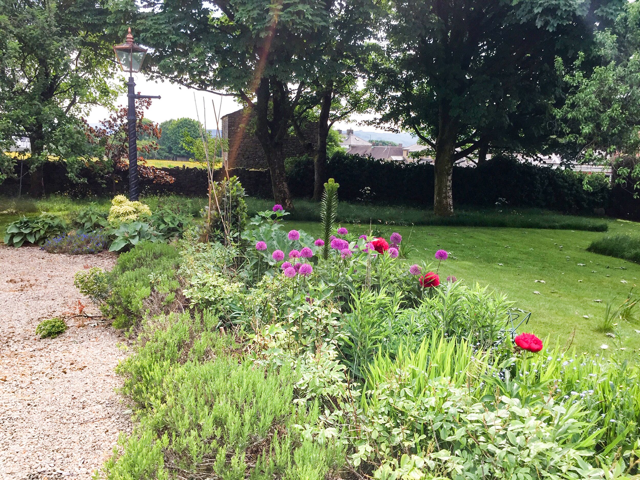A garden with various plants and flowers, a gravel path, and a brick building in the background.