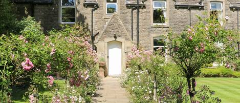 A stone house with a central door, flanked by flower beds and a paved pathway leading to the entrance.