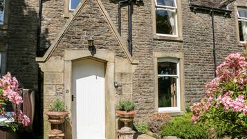 A stone house with a white door, multiple windows, and a small garden with pink flowers.