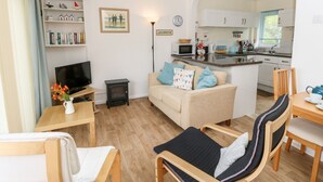 A compact living space with a sofa, a TV, a small table, a bookshelf, and a kitchen area visible through an archway. - Buddleia Cottage (Seaview)