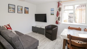 Een moderne woonkamer met een grijze bank, een donkerblauwe fauteuil, een eettafel met stoelen, een flatscreen televisie aan de wand en een raam met gordijnen met een patroon.