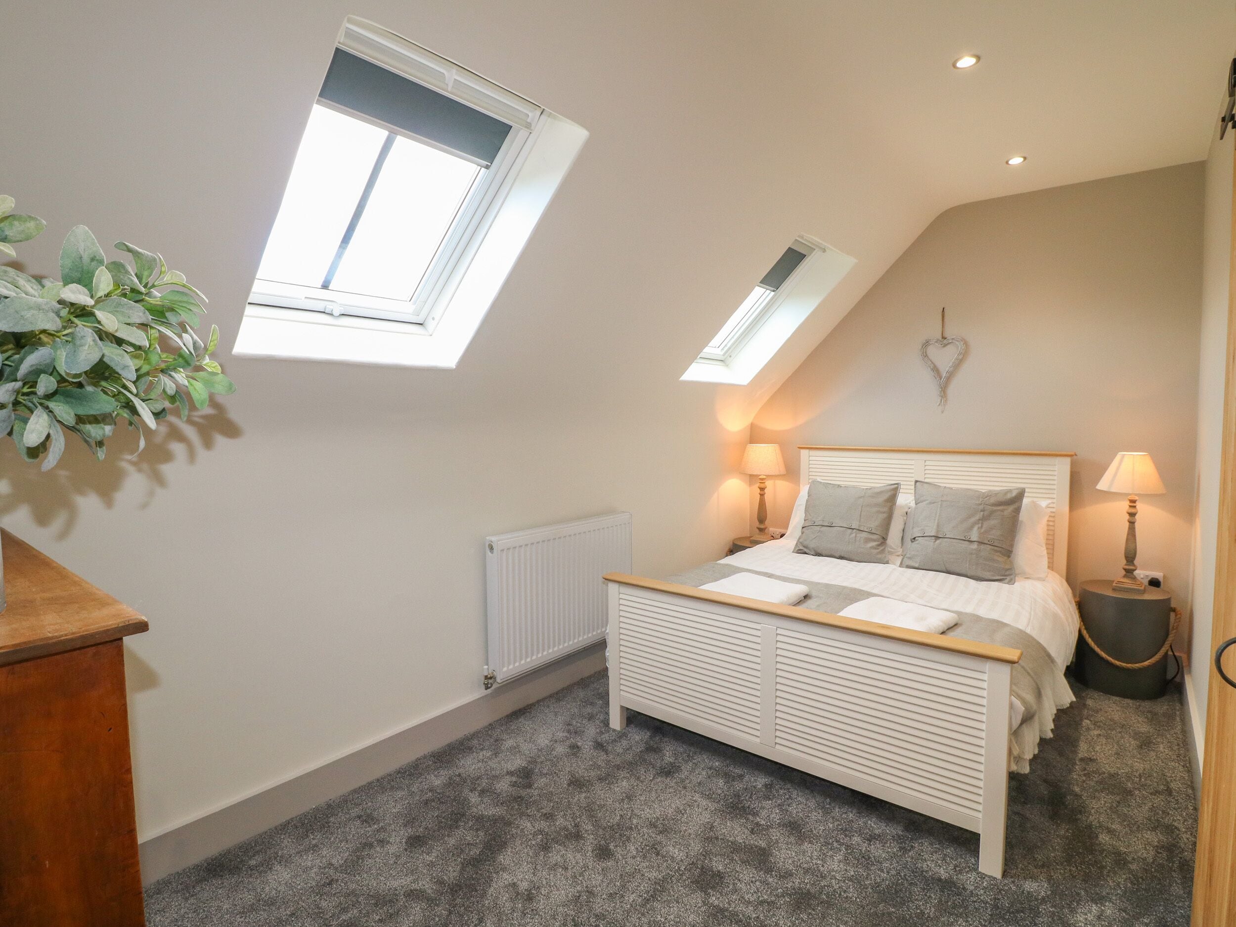 A bedroom with a sloped ceiling, a skylight, a radiator, a bed with pillows, a bedside table with a lamp, and a plant.