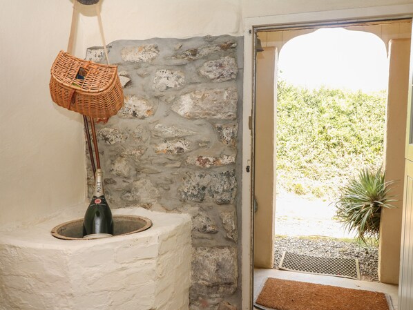 A stone wall, a wicker basket, a bottle, and a door leading to an outdoor area with a plant. - Higher Roskorwell Cottage (Helston)