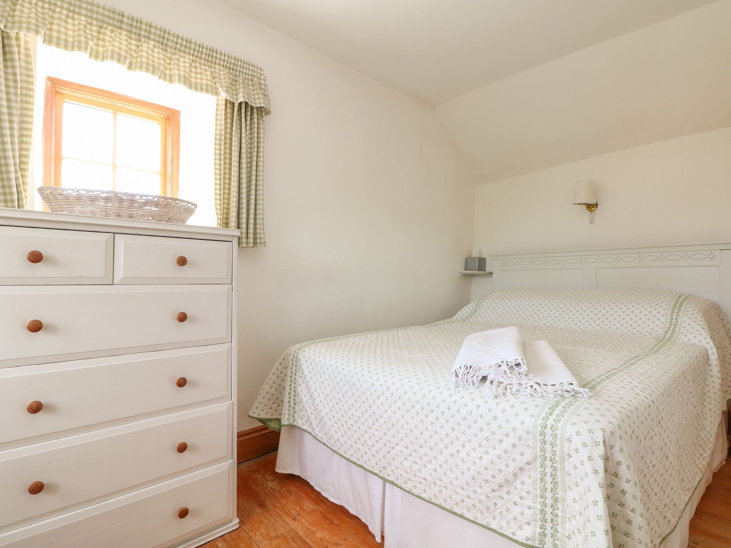 Un dormitorio con cama, cómoda con cajones, ventana con cortinas y una lámpara de pared.