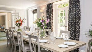 Een eetkamer met een lange tafel gedekt voor zes personen, omringd door zes stoelen. Er staan bloemstukken op de tafel, en er is een raam met behang gordijnen te zien.